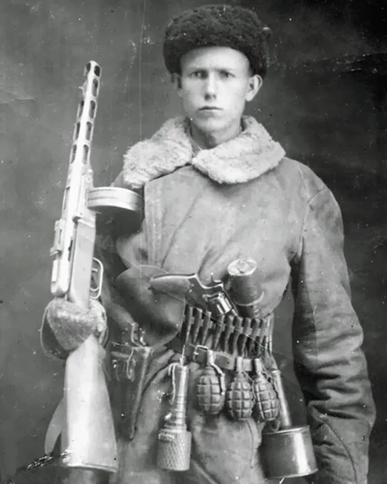 A person wearing a fur hat and coat holds a PPSH-41 submachine gun. The person has a belt with various grenades and equipment attached. The style and items suggest a mid-20th-century military setting.