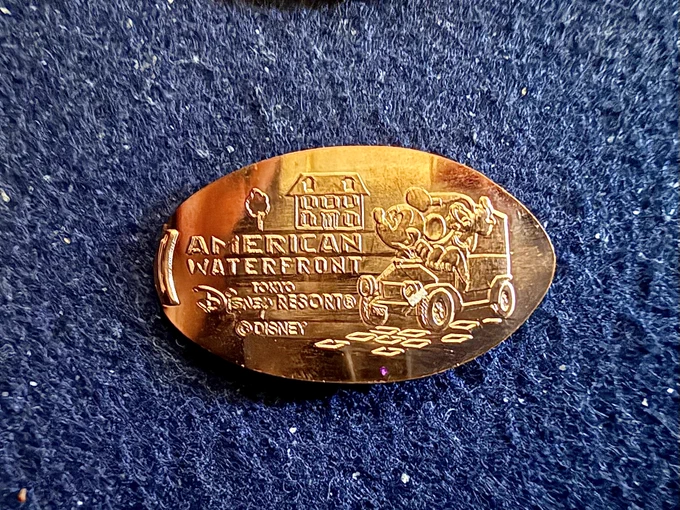 Souvenir medal ("pressed penny" of Mickey and Pluto driving a vintage delivery truck on cobblestone with a home in the background. Text: "American Waterfront" "Tokyo Disney Resort" ©Disney
