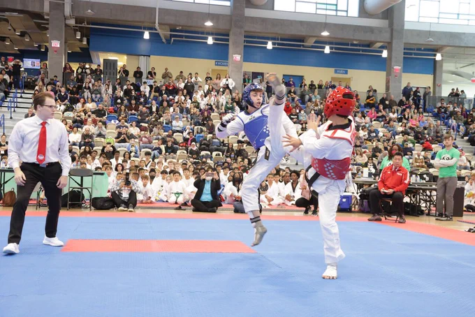 Two athletes in protective gear competing in a martial arts match on mats, with a referee watching and a crowd in the background.
