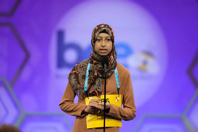 A contestant at the Scripps National Spelling Bee stands at the microphone on stage, wearing a patterned headscarf and holding a yellow identification badge.