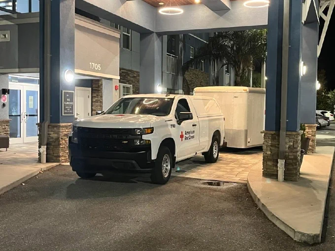 A white truck with an "American Red Cross" logo is parked under a building entrance canopy. The truck is attached to a white trailer. Nearby, there are illuminated entrance signs and decorative pillars.