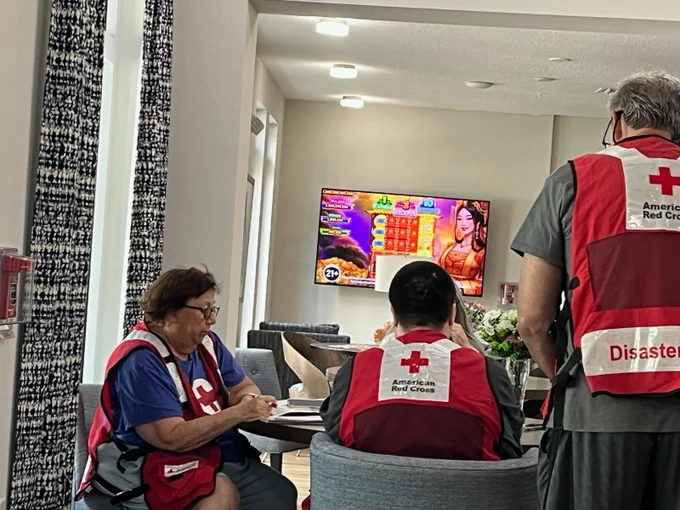 Red Cross volunteers are seated around a table in a lounge area, wearing vests with the "American Red Cross" logo. A television in the background displays a bright game show with Indian design elements.
