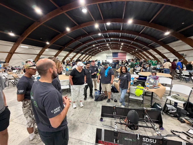 A group of individuals stands around a large drone inside an arched hangar. The machine has "SDSU" visibly marked on its parts. In the background, there are tables with equipment and an American flag hanging on the wall.
