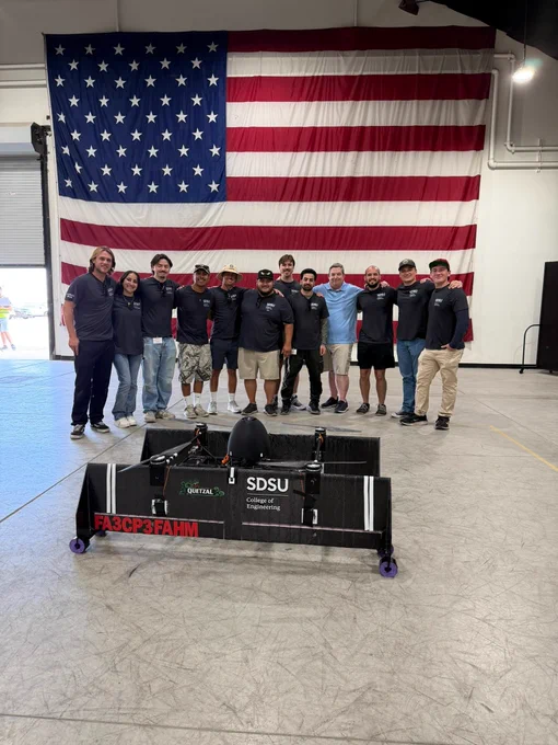 A group of people wearing matching shirts stands indoors in front of a large American flag. In front of them is a device labeled "SDSU."