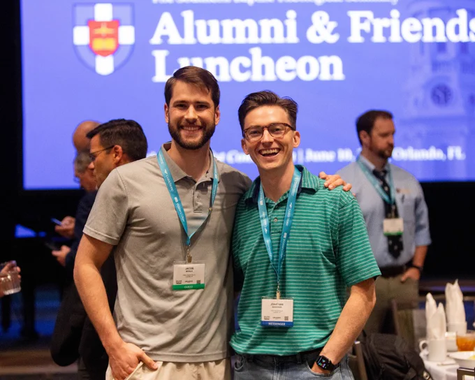 Two attendees at an Alumni & Friends Luncheon smile for the camera. The event is lively and well-attended.