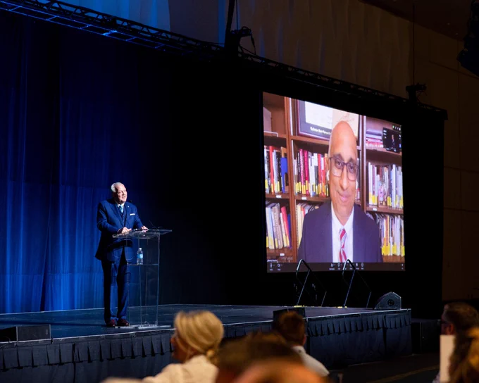 A presenter speaks onstage at a conference with a large video call screen displaying another speaker. The setting is professional, and the mood is formal.