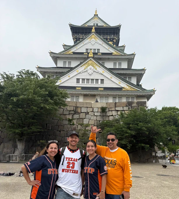 A group of four people standing in front of Osaka Castle. Three are wearing UTEP Miner jerseys, one is wearing a Texas Western jersey. They are smiling and posing for the photo.
