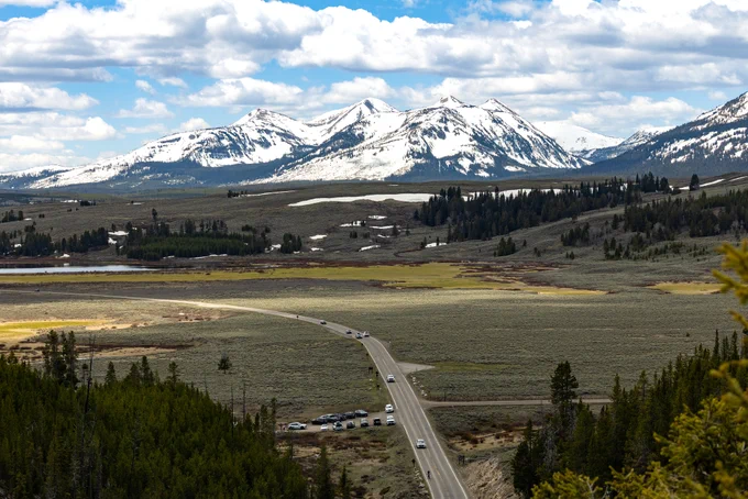An aerial view of cars on a paved road that winds through a mountain valley. Snow-capped mountains can be seen in the distance.
