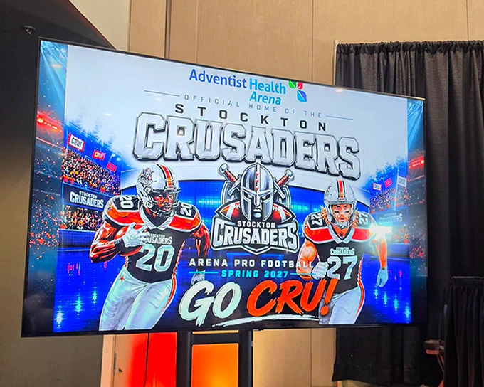A promotional screen displays an advertisement for "Stockton Crusaders," an arena pro football team for Spring 2027. The image includes logos and two football players in uniforms. The background shows a stadium filled with cheering crowds. The top of the screen features the "Adventist Health Arena" logo.