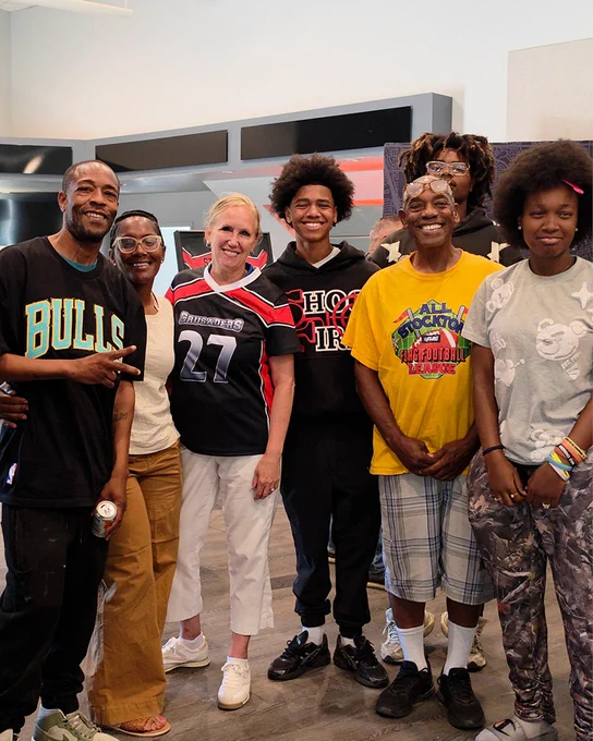 A group of people are standing together with Stockton Mayor Christina Fugazi smiling. One person is wearing a Chicago Bulls shirt, and another is wearing a "Chicago 27" sports jersey. The background shows a modern interior with a dark countertop and some decor.