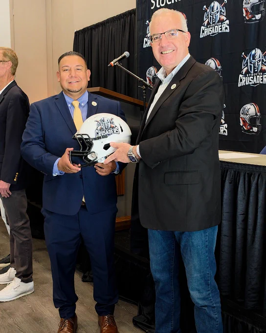 Stockton Councilmember Brando Villapudua and Councilmember Michael Blower smiling and holding a Stockton Crusader football helmet. The background has a banner with the Stockton Crusaders logo.