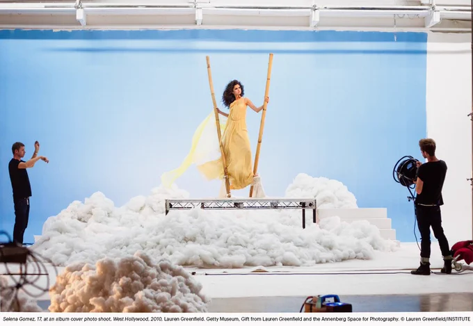 Color image of a staged photoshoot set with a blue backdrop and artificial clouds, showing Selena Gomez in a flowing yellow dress standing on a raised platform, while crew members and lighting equipment are visible on either side.