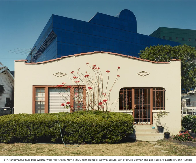 Small cream-colored single-story house with a curved roofline, red trim, and barred front door, set behind a hedge and flowering plants; a large blue modern building rises prominently in the background.