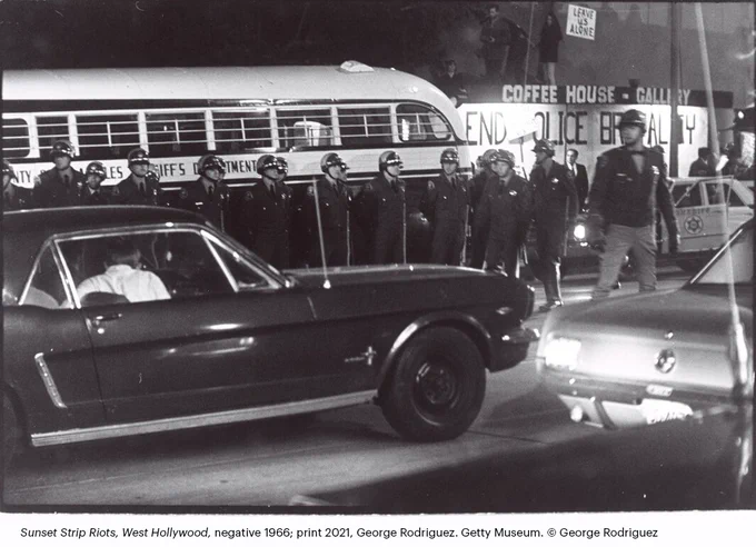 Black-and-white nighttime street scene with a line of uniformed police officers standing in formation beside a bus, while cars pass in the foreground and signs in the background read “End Police Brutality.” 
