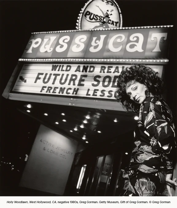 Black-and-white nighttime street scene showing a person with big, loose curls and hoop earrings standing beneath a brightly lit theater marquee reading “Pussycat” and “Future Shock / French Lessons.”