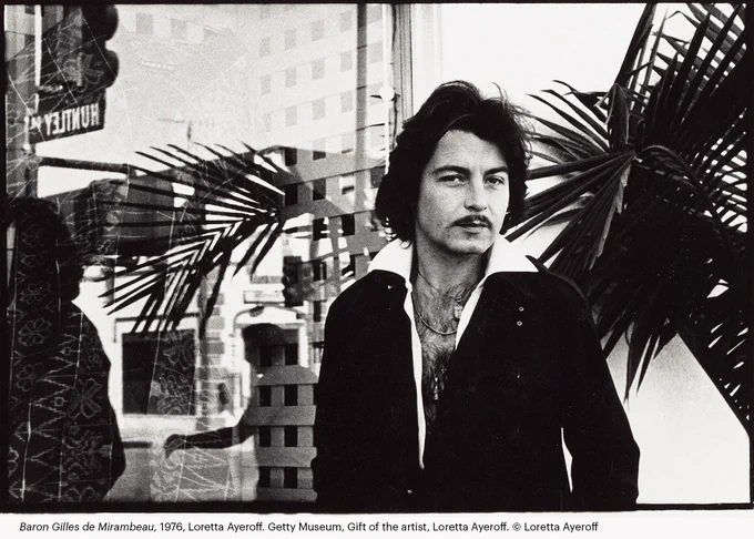 Black-and-white photograph of a person standing indoors near large windows and leafy plants, wearing a dark open-collared shirt, with layered reflections of West Hollywood visible in the glass behind.