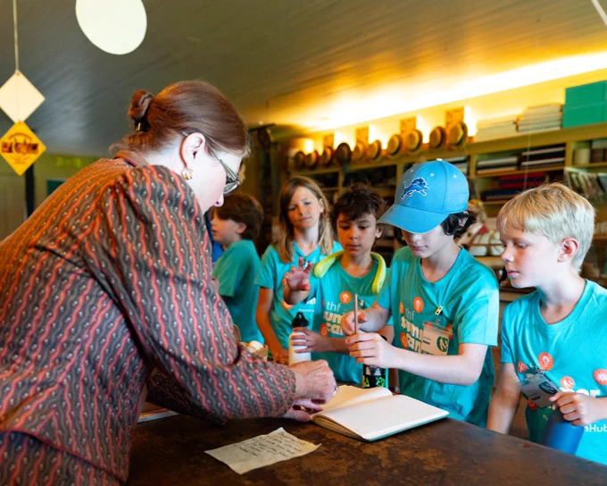 A person in a vintage dress is pointing toward a book at a table. Nearby, a group of children are observing. The setting has shelves and lighting in the background.