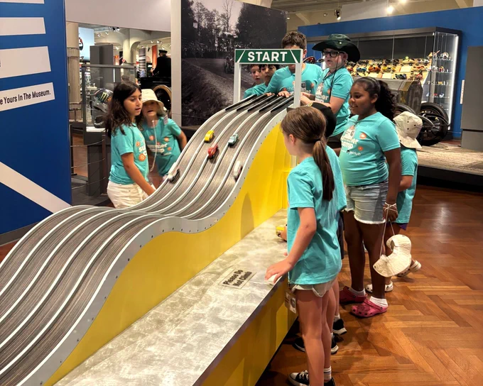 A group of children wearing matching blue shirts, are gathered around a slot car race track with four lanes. Small colorful cars are set on the track, which starts under a "START" sign. In the background, there are museum exhibits and displays on shelves.