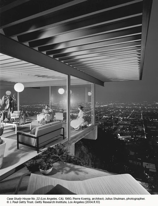 Black-and-white photograph of a glass-walled modern home with two seated figures inside under round ceiling lights, overlooking a sprawling cityscape of glowing lights below.
