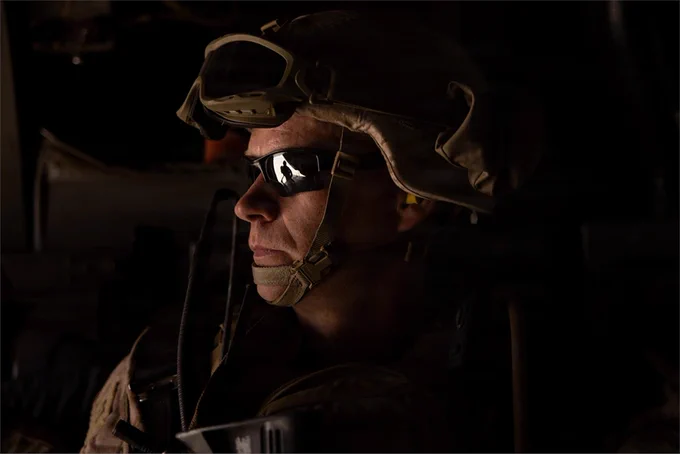 A Marine wearing a helmet and protective eyewear sits inside an aircraft, with another Marine reflected in his sunglasses.