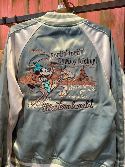 The back of a light blue and white satin bomber style jacket with spiraling buttes, Mickey riding a horse with a lasso in his hand with "Rootin' tootin' Cowboy Mickey! He's a hero for all the folks in Westernland" embroidered on the back.