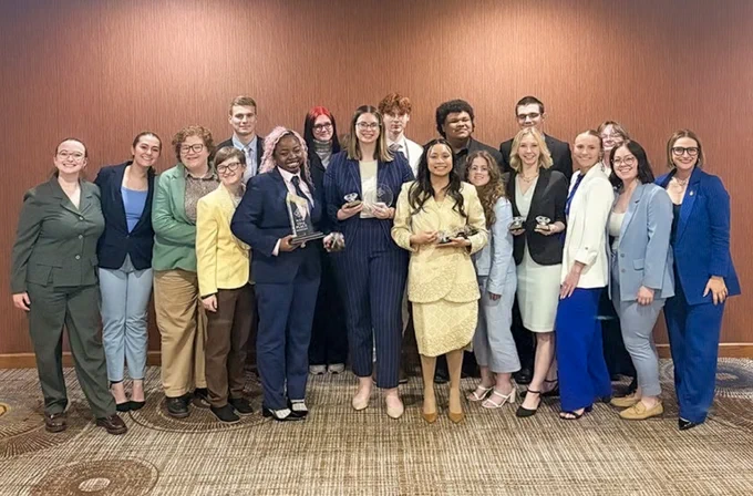 A group of people standing in a room, dressed in formal attire, holding awards. The background features a brown wall and patterned carpet.