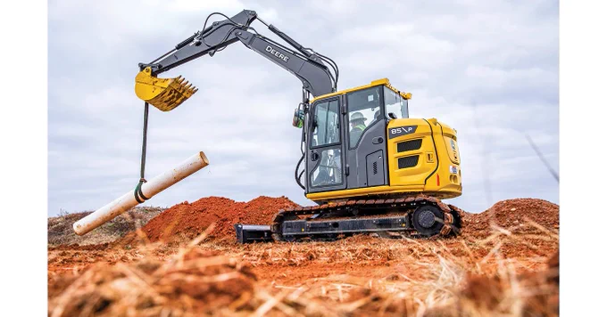 A yellow John Deere excavator is lifting a long white pipe on a construction site with reddish soil. The sky is overcast.