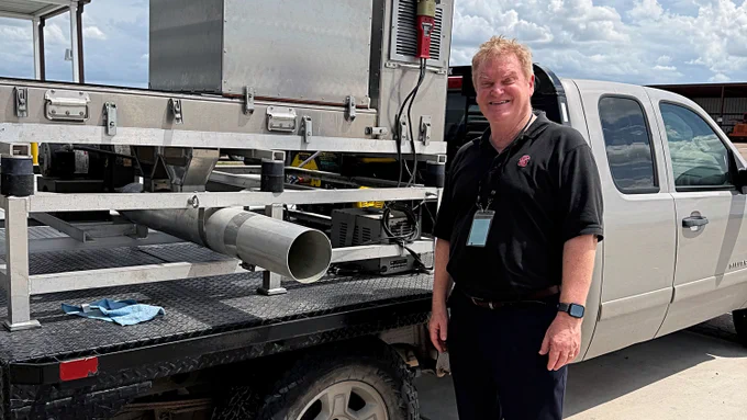 Man in black polo and badge standing next to a white pickup truck with industrial equipment mounted on the truck bed under a partly cloudy sky.