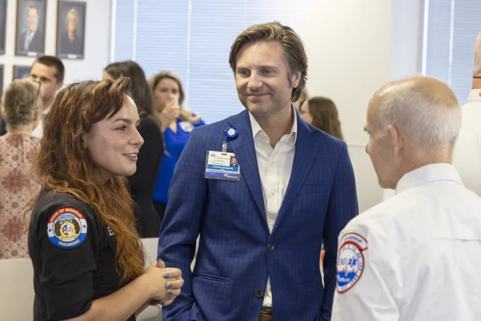 Three individuals converse in a bright room. A woman in a black top with a \"STATE OF MISSOURI PARAMEDIC\" patch talks to Andrew Hedgpeth, Cox Health VP of Human Resources, who wears a blue suit and a name badge. An older man in a white EMS uniform stands to the right.