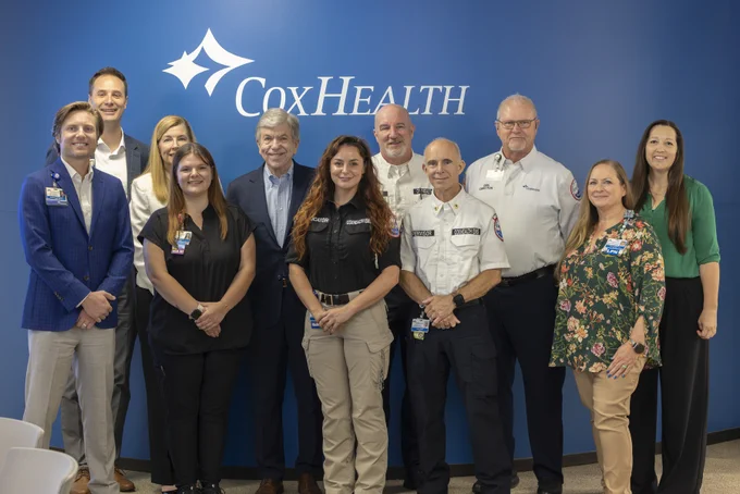Twelve CoxHealth professionals, including men in suits & EMS uniforms, and women in business casual & scrubs, smile in a group photo against a blue wall with the CoxHealth logo. Titles like MA III, EDUCATOR, PERVISOR, EMS DIRECTOR, and LPN are visible on their badges.