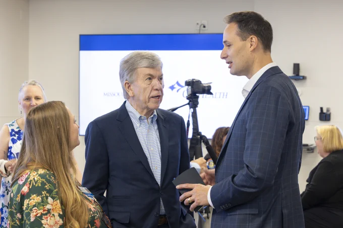 Older man in a blue suit talking to a younger man in a plaid suit holding a notebook. A woman in a floral top listens intently. In the background, a screen displays \"MISSOURI HEALTH\" with a logo and a camera on a tripod. Other attendees are partially visible.