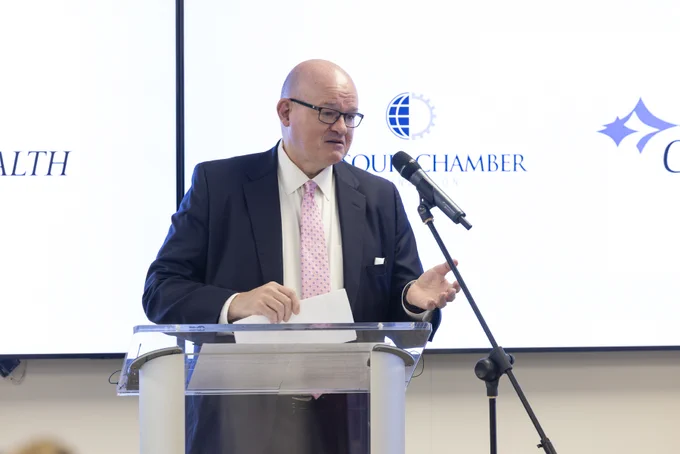 A man in glasses, a dark suit, and pink tie speaks from a clear podium, holding papers and gesturing. A microphone is in front of him. Behind, a screen shows a globe/gear logo with 'OUR CHAMBER' and partial text 'ALTH' and another blue logo.