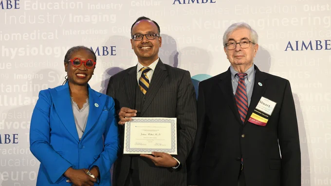 Three individuals are standing together at an AIMBE event. The person in the center is holding a certificate. The background displays words related to biomedical engineering.