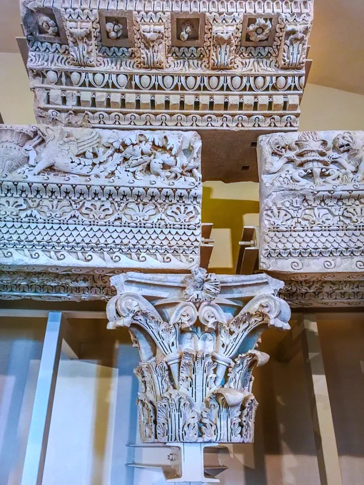 ​RABIRIUS.
DECORATIVE CARVING OF THE SUMMA CAVEA OF DOMITIAN'S THEATRE, LATE C1 CE. VILLA PONTIFICIA DI CASTEL GANDOLFO

Here we see a reconstruction of disparate pieces of a colonnade from the theatre in Domitian's villa, the Albanum. An ornate column capital supports a densely-carved entablature. The horizontal beams feature bands of repeating leaf motifs, delicate geometric patterns, and a rich figurative frieze where a winged griffin dissolves into swirling vines. Above, the lintel bears the architect Rabirius's signature, a unique double-ring motif carved into the spaces between the rectangular dentils. This hyper-detailed Flavian Baroque style rejected older, simpler designs to maximise dramatic chiaroscuro under the Italian sun, projecting raw imperial power. Yet this lace-like stonecarving formed a gilded cage, framing a court defined by psychological dread where the paranoid, universally reviled emperor Domitian terrified his ruling class. 