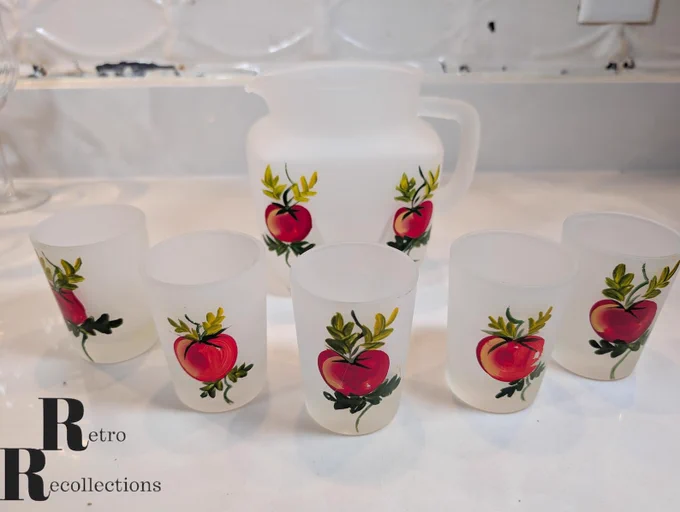 Hazel Atlas frosted glass pitcher and five matching juice glasses hand‑painted by Gay Fad Studios with red apples and green leaves, arranged on a white countertop
