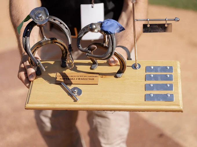 A person is holding a wooden trophy featuring metal sculptures shaped like abstract figures made of horseshoes. An affixed plaque reads "Softball Challenge.