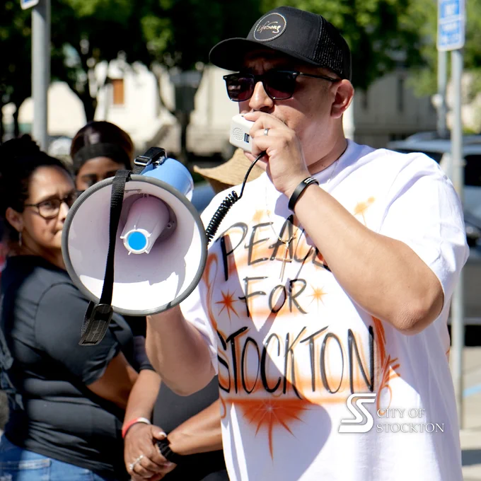A person with sunglasses and a cap speaks into a megaphone, wearing a shirt that reads "Peace for Stockton." A group of individuals stands in the background. A "City of Stockton" watermark is visible in the corner.