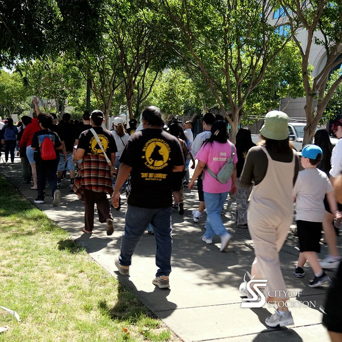 A group of people is marching on a sidewalk under trees, wearing casual clothing. Some have shirts with logos, and one reads "City of Stockton." The atmosphere appears lively and communal.