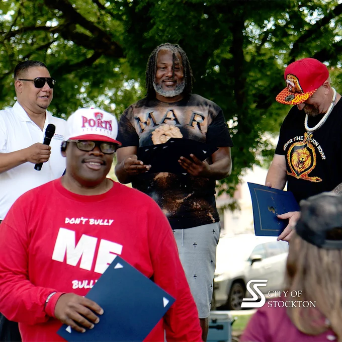 Councilmember Brando Villapudua and three other people are seen outdoors holding certificates, with lush green trees in the background. One person is speaking into a microphone and wearing sunglasses, another is wearing a "Don't Bully Me" shirt and a Ports baseball cap. The City of Stockton logo is visible in the corner.