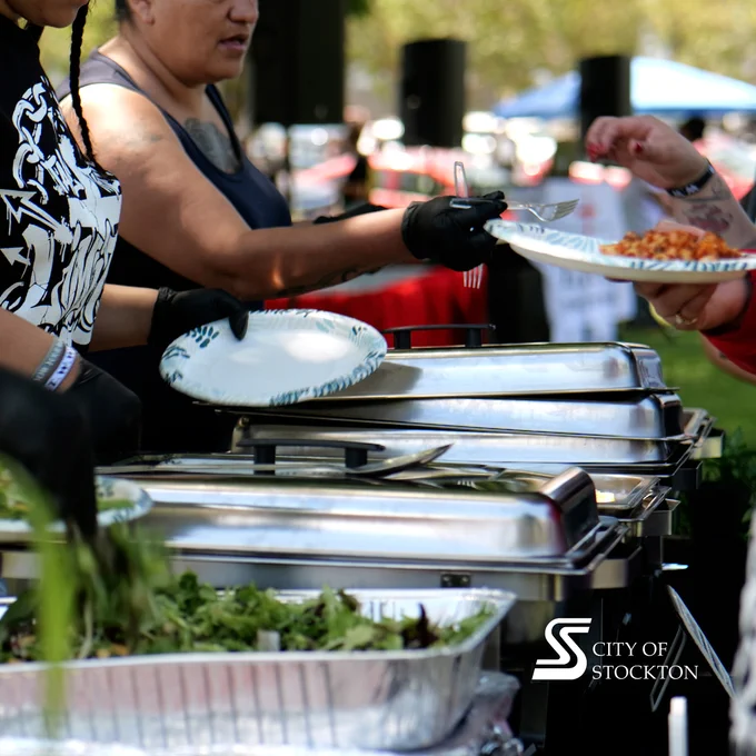 People are serving food from a buffet setup at an outdoor event. Chafing dishes are filled with various foods, and one person holds a plate while being served. The logo of the City of Stockton is visible in the corner.