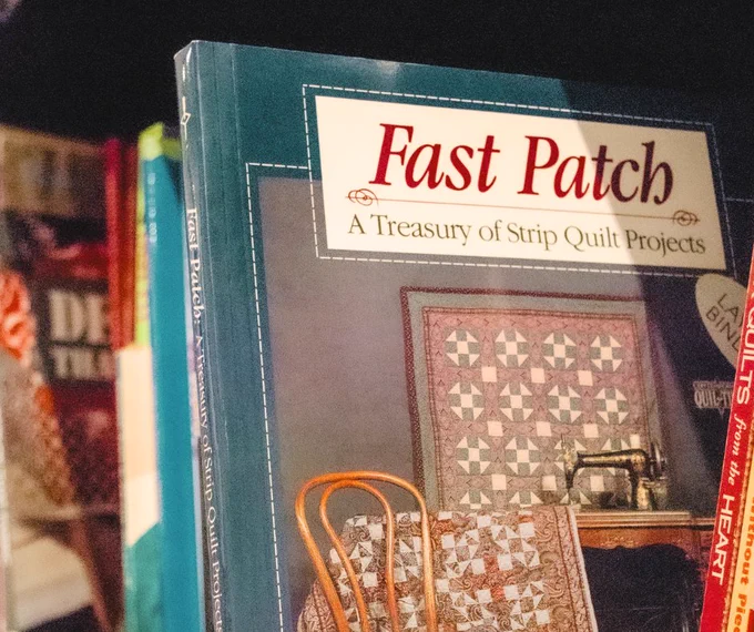 A bookshelf containing various quilting books. A prominently featured book titled "Fast Patch: A Treasury of Strip Quilt Projects" is visible, showcasing a chair and quilts on its cover. Other books are partially visible on the shelf.