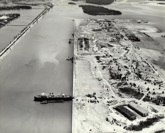 Aerial black and white photo showing a large body of water on the left with a long causeway lined with trees. On the right, a vast land area under development with dirt roads, scattered trees, and several buildings. A large ship and smaller boat are docked at the land's edge.