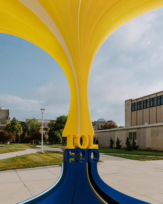 A colorful, abstract sculpture featuring large yellow and blue curves is situated outdoors on a university campus. The sculpture prominently displays the word "TOLEDO" in bold letters. 