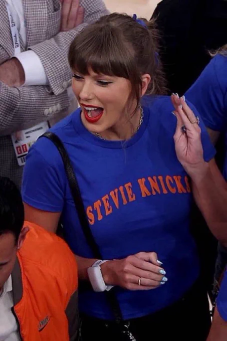 taylor swift at a knicks game wearing a blue shirt that reads “stevie knicks” (spelled like the team) and an arena light-up bracelet. she’s looking to the side waving at someone with an open mouth smile.