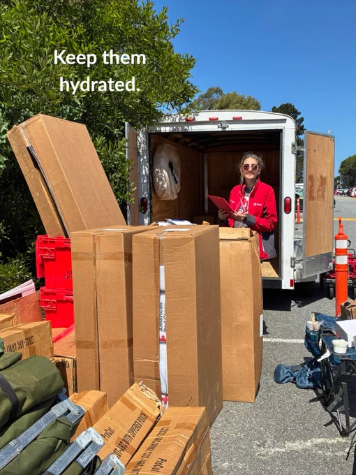 A person stands inside an open trailer surrounded by tall cardboard boxes and equipment. They are holding a clipboard. The text "Keep them hydrated" is visible at the top left. The scene takes place outdoors on a clear, sunny day.