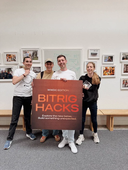 Alt text: A group of four smiling people standing together indoors, celebrating their achievement. They are holding a large brown square sign that reads "WWDC EDITION BITRIG HACKS" in bold orange text, with the subtitle "Explore the new betas. Build something unexpected." One person on the right is holding up a small glass cube trophy. The wall behind them is decorated with framed pictures and a whiteboard with diagrams.