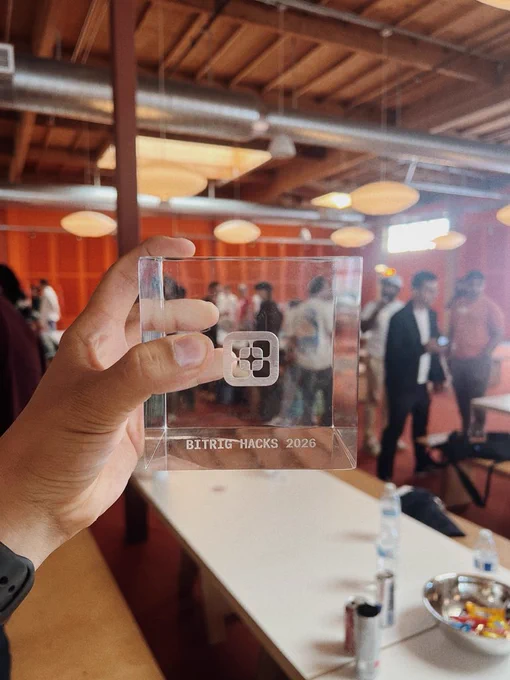 A close-up shot of a hand holding a clear, square glass trophy. The trophy is engraved with a square geometric logo in the center and the text "BITRIG HACKS 2026" near the bottom. The background is a softly blurred, brightly lit indoor event space with wooden ceilings, tables, and people mingling.