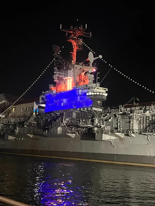 A decorated battleship is illuminated with orange, blue, and white lights at night. The ship is moored in calm water, with strings of white lights outlining its structure.