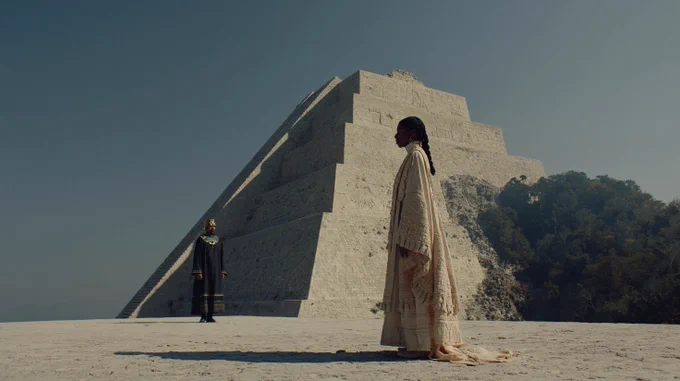 A cinematic still in the visual style of Denis Villeneuve, photographed with the naturalistic lighting of Greig Fraser on a large-format Alexa LF sensor. A vast stone platform at the top of a Mayan pyramid stretches into open sky, carved glyph patterns embedded in the surface. A female emissary stands centre-right, facing left, still and composed, with Indigenous Mesoamerican features, copper-brown skin with warm golden undertones, wearing layered ceremonial robes with jade and carved stone ornamentation. Opposite her stands a man of status centre-left, dressed in dark ceremonial garments with gold and jade detailing. The space between them is wide and deliberate. A sharp band of sunlight cuts across the platform, dividing light and shadow, with deep negative fill shaping both figures. Warm desaturated tones, subtle haze, strong symmetry, grounded realism, film grain.