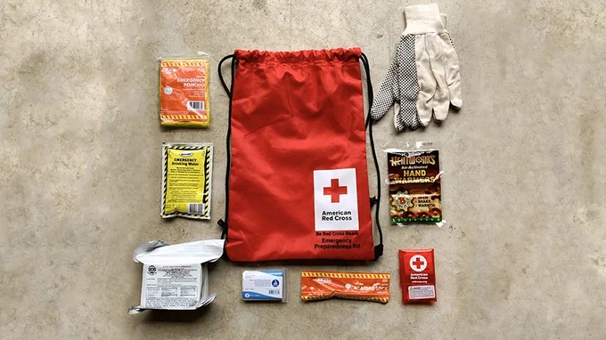 A red emergency preparedness kit bag from the American Red Cross is centered on a concrete surface. Surrounding it are various items, including food and water packets, gloves, a small flashlight, and a first aid kit.